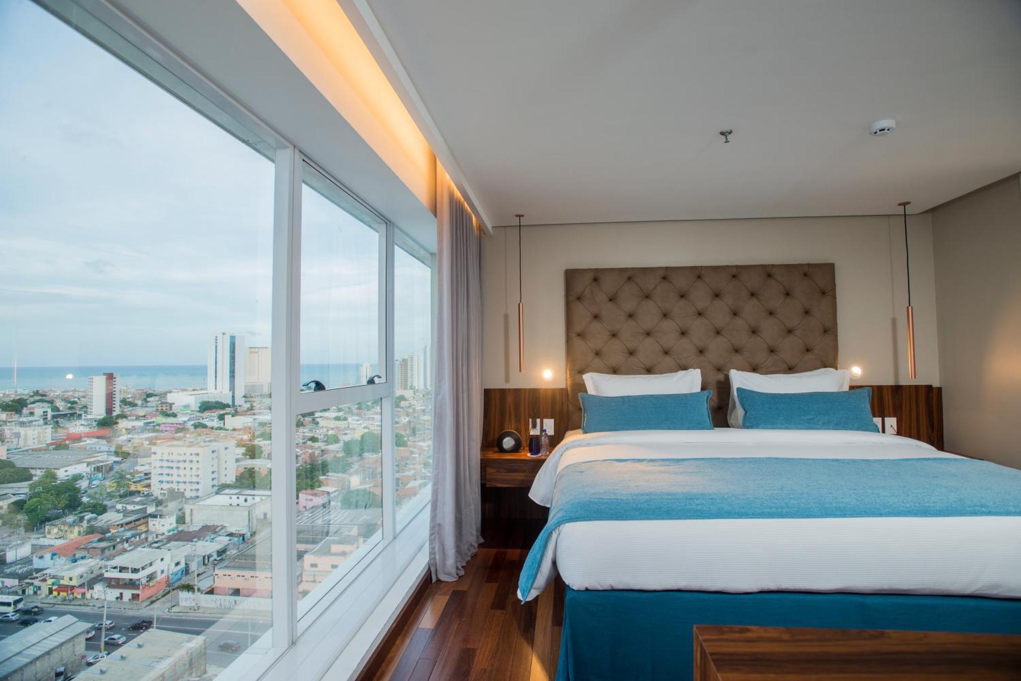 Hotel Luzeiros Recife Bagian luar foto The photo shows a modern hotel room featuring a large window with a city view. Inside, there are two neatly made beds with light blue and white bedding, flanked by wooden nightstands. The room has a contemporary design, with a padded headboard and so
